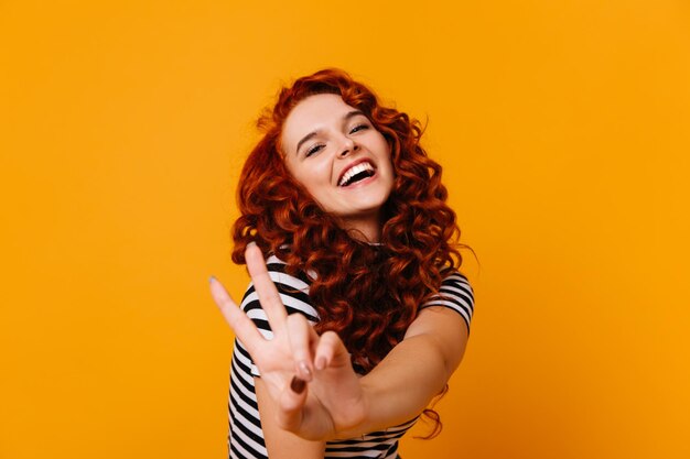 Joven activa con el pelo rojo se ríe y muestra el signo de la paz en el estudio naranja