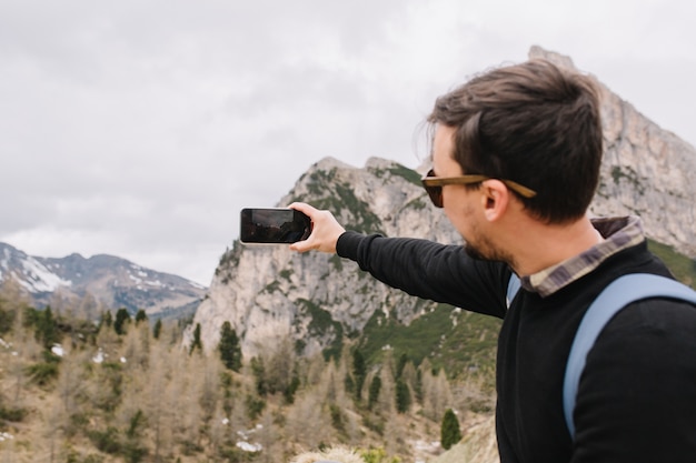 Joven activo con cabello castaño oscuro con camisa debajo de un suéter negro viajando en las montañas y video grabado en el teléfono inteligente