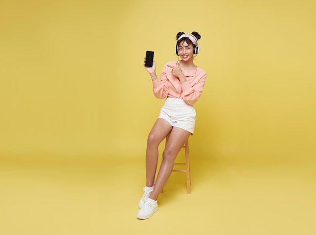 Una joven adolescente asiática señala con la mano un teléfono inteligente mientras escucha música con auriculares y se sienta en una silla aislada en un fondo amarillo