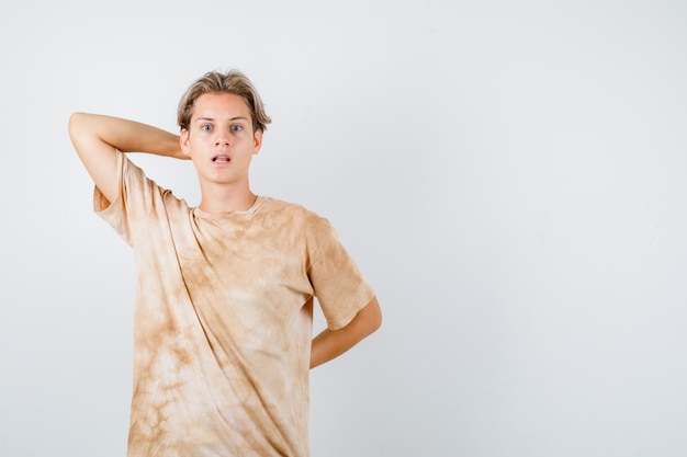 Foto gratuita joven adolescente en camiseta manteniendo la mano detrás de la cabeza y la espalda y mirando desconcertado, vista frontal.