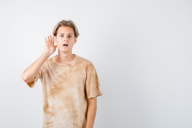 Joven adolescente en camiseta manteniendo la mano detrás de la oreja y mirando perplejo, vista frontal.