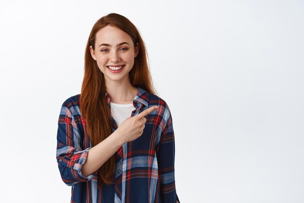 Joven adolescente natural con el pelo largo y rojo señalando con el dedo en la esquina superior derecha mostrando el logotipo de texto promocional sonriendo dientes blancos de pie sobre fondo blanco