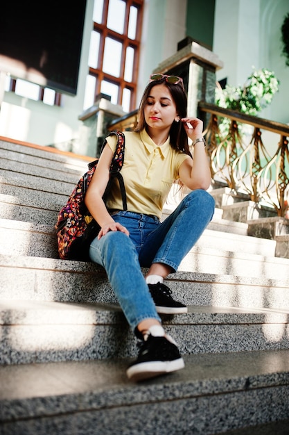 Joven adolescente sentada en las escaleras vestida con camiseta amarilla, jeans y gafas de sol con mochila