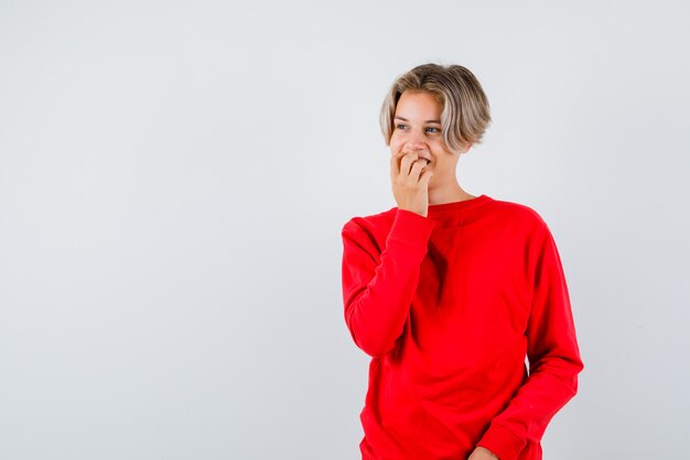 Joven adolescente en suéter rojo mordiéndose las uñas mientras mira a otro lado y parece feliz, vista frontal.