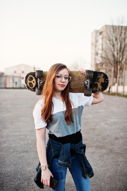 Joven adolescente urbana con monopatín en gorra de gafas y jeans rasgados en el campo de deportes del patio al atardecer haciendo selfie en el teléfono