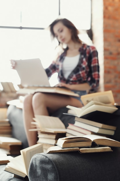 Joven adolescente usando la computadora portátil rodeada de muchos libros.