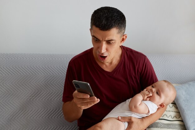 Joven adulto sorprendido con camiseta marrón de estilo casual sosteniendo a una niña o un niño en brazos y usando el teléfono móvil mientras está sentado en el sofá en la sala de estar, recibe noticias impactantes por correo electrónico.