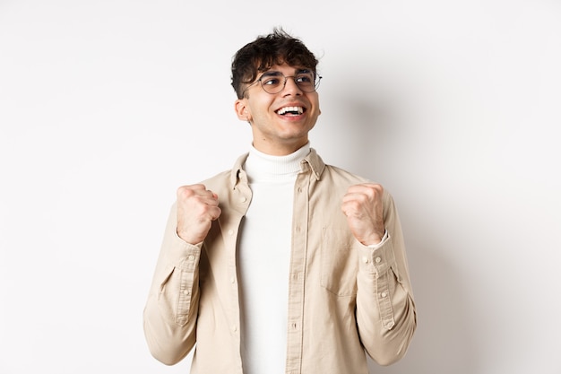 El joven afortunado dice que sí con cara de alivio, sonriendo y haciendo puñetazos, triunfando del éxito, ganando el premio, de pie sobre un fondo blanco después de lograr el objetivo