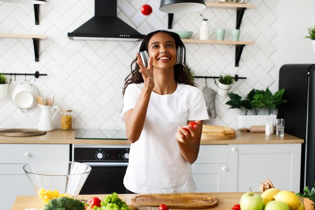 Joven africana está escuchando música en los auriculares y está haciendo malabarismos con tomates cherry en la cocina