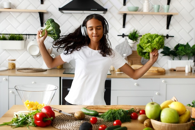 Joven africana está feliz escuchando música a través de auriculares con los ojos cerrados y sostiene un brócoli y ensalada