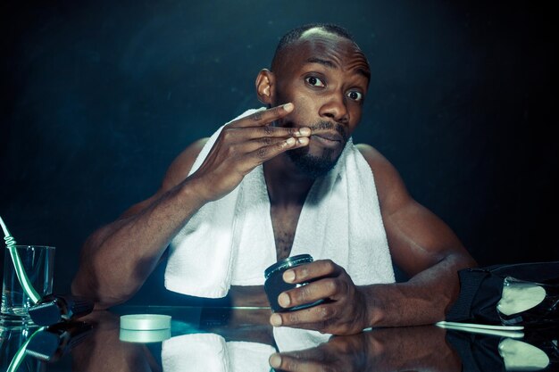 El joven africano en el dormitorio sentado frente al espejo después de rascarse la barba en casa. Concepto de emociones humanas. conceptos de crema para después del afeitado