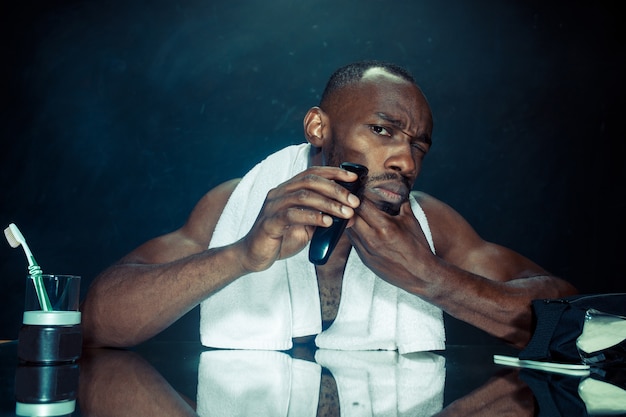 Foto gratuita joven africano en el dormitorio sentado frente al espejo rascándose la barba en casa