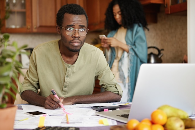 Joven africano sintiéndose estresado pagando facturas en línea, calculando gastos de gas y electricidad, sentado en la mesa de la cocina frente a la computadora portátil abierta y tomando notas. Estrés financiero y deudas