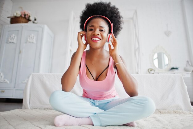 Joven afroamericana en camiseta rosa escucha música con auriculares en su habitación blanca