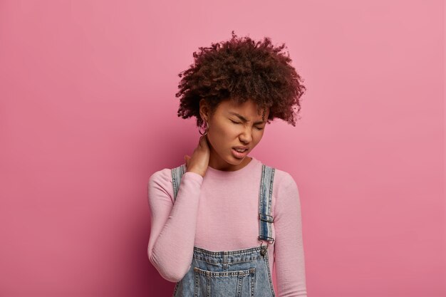 La joven afroamericana disgustada siente malestar en la columna vertebral, se toca el cuello y frunce el ceño por el dolor, lleva un estilo de vida sedentario, se viste de manera informal, posa contra la pared rosa pastel, es fatiga