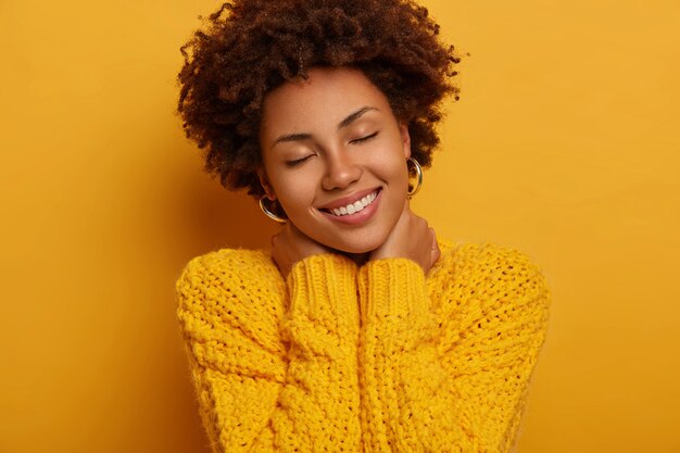 joven afroamericana rizada mantiene los ojos cerrados, toca el cuello con ambas manos, inclina la cabeza, viste un suéter amarillo de punto, posa en interiores.