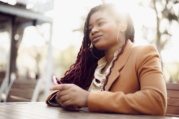 Joven afroamericana sentada en la cafetería de la calle y sosteniendo el teléfono inteligente en sus manos