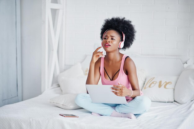 Joven afroamericana sentada en la cama mientras trabaja en una computadora portátil y escucha música en los auriculares