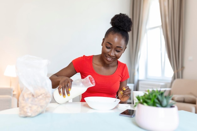 Una joven afroamericana sonriente vierte copos de maíz en un plato con leche La niña tiene un desayuno saludable en una casa elegante y acogedora por la mañana mientras revisa su correo electrónico en una laptop