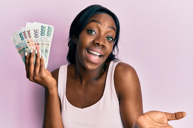 Joven afroamericana sosteniendo billetes de corona checa celebrando el logro con una sonrisa feliz y expresión ganadora con la mano levantada