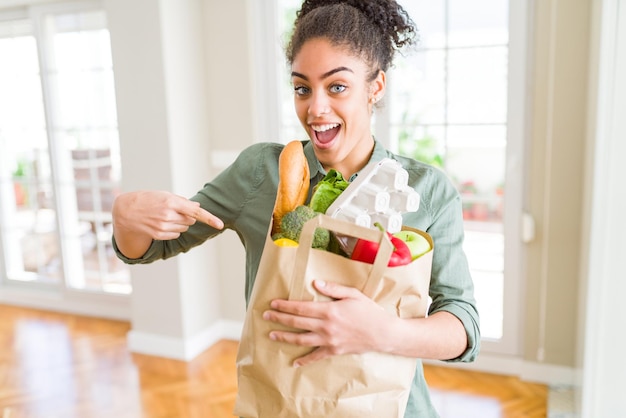 Joven afroamericana sosteniendo una bolsa de papel de comestibles del supermercado muy feliz señalando con la mano y el dedo