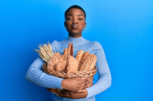 Foto gratuita joven afroamericana sosteniendo una canasta de mimbre con pan hinchando las mejillas con cara graciosa boca inflada con aire atrapando aire