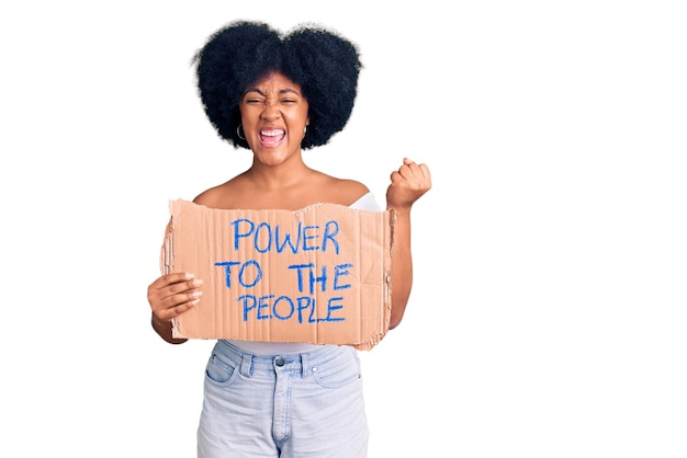 Joven afroamericana sosteniendo el poder de la pancarta de la gente gritando orgullosa, celebrando la victoria y el éxito muy emocionada con los brazos levantados