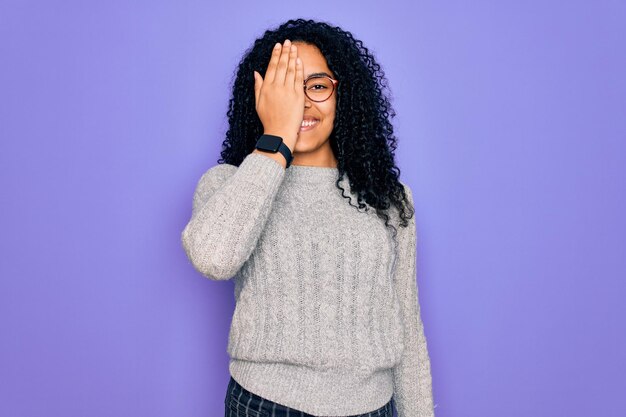 Foto gratuita joven afroamericana con suéter casual y anteojos sobre fondo morado que cubre un ojo con una sonrisa segura en la cara y emoción sorpresa