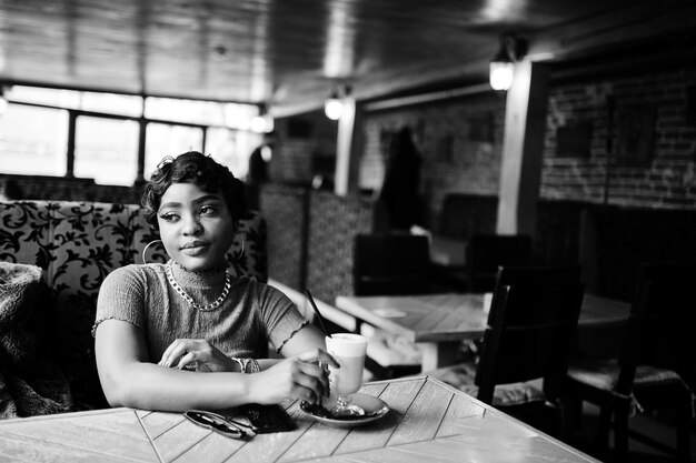 Joven afroamericana urbana con top rosa sentada en un café y bebiendo café con leche