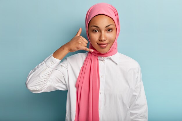 Joven afroamericana usa hijab, hace un gesto de llamarme, da forma a las manos como un teléfono cerca de la oreja, tiene una expresión facial satisfecha, habla a distancia con alguien