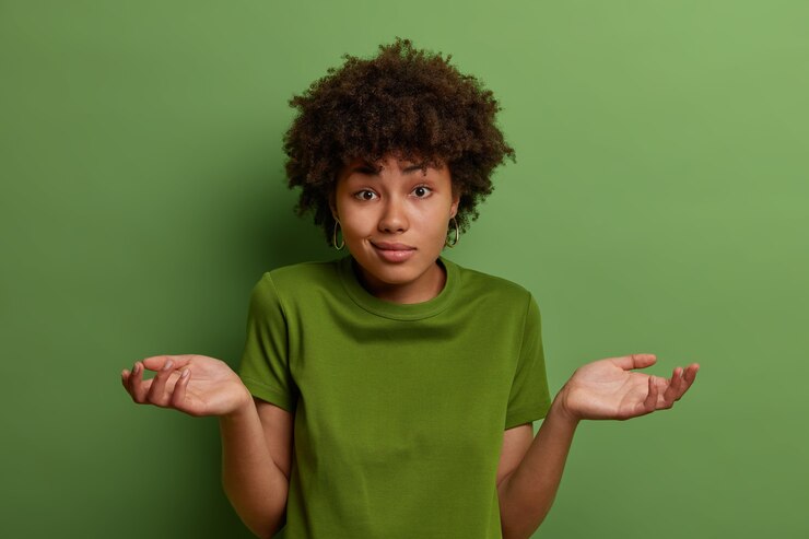 Foto gratuito la joven afroamericana vacilante y desconcertada extiende las palmas de las manos, se siente insegura, se ve desconcertada, se pone de pie, usa una camiseta verde casual en un color con fondo, toma una decisión difícil