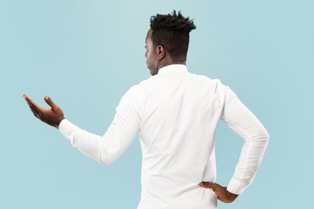 Joven afroamericano aislado sobre fondo azul de estudio, expresión corporal. Hermoso retrato masculino de media longitud fotografiado desde atrás. Concepto de emociones humanas, expresión facial.