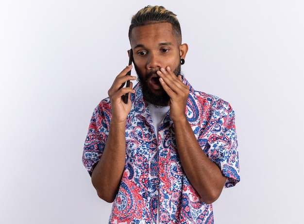 Joven afroamericano en camisa colorida mirando asombrado y conmocionado mientras habla por teléfono móvil de pie sobre blanco
