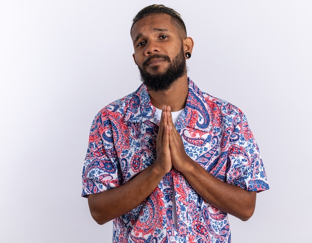 Foto gratuita joven afroamericano en camisa colorida mirando a la cámara con expresión de esperanza sosteniendo las palmas juntas