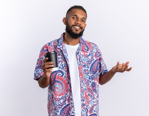 Joven afroamericano en camisa colorida mirando a la cámara sonriendo alegremente sosteniendo la taza de papel de pie sobre blanco