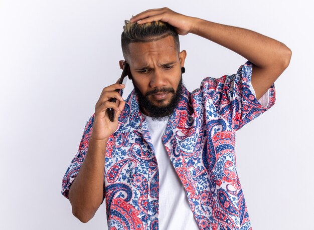 Joven afroamericano en camisa colorida mirando confundido mientras habla por teléfono móvil de pie sobre blanco