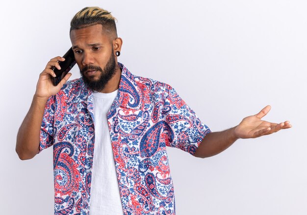 Joven afroamericano en camisa colorida mirando confundido mientras habla por teléfono móvil de pie sobre fondo blanco.