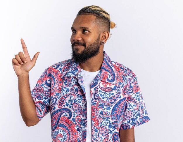 Joven afroamericano en camisa colorida mirando a un lado sonriendo alegremente apuntando con el dedo índice hacia el lado de pie sobre fondo blanco.