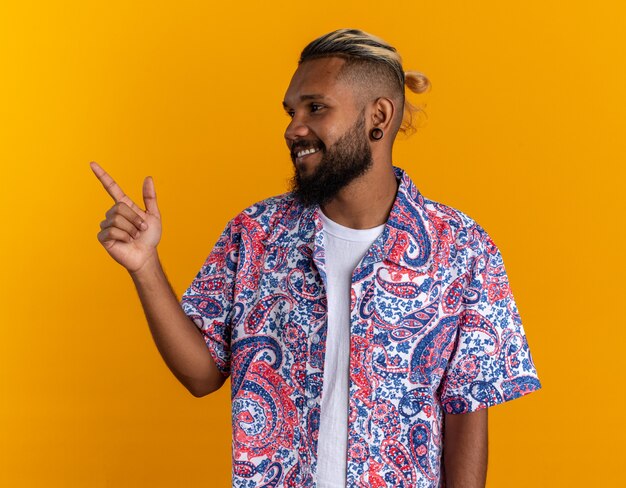 Joven afroamericano en camisa colorida mirando a un lado sonriendo alegremente pointng con el dedo índice hacia el lado de pie sobre fondo naranja
