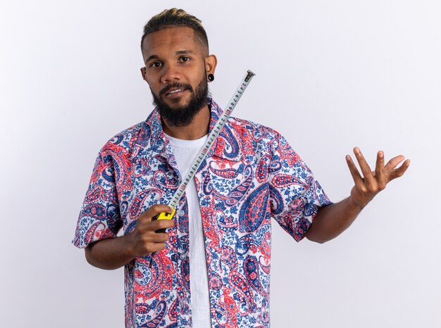 Joven afroamericano en camisa colorida sosteniendo cinta métrica mirando a la cámara sonriendo con cara feliz de pie sobre fondo blanco.