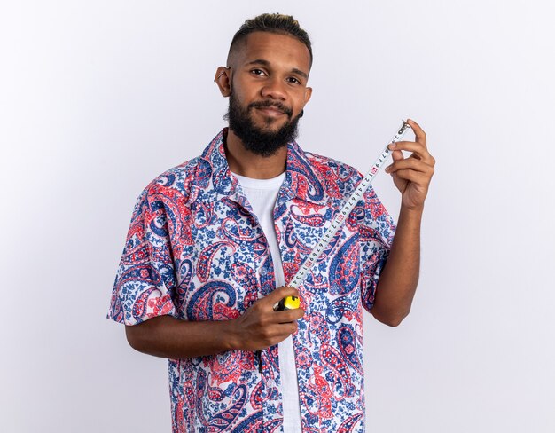 Joven afroamericano en camisa colorida sosteniendo cinta métrica mirando a la cámara con una sonrisa en la cara feliz y positiva de pie sobre fondo blanco.