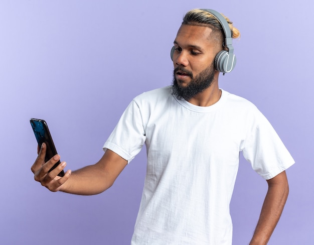 Joven afroamericano en camiseta blanca con auriculares mirando la pantalla de su teléfono inteligente con una sonrisa en la cara de pie sobre fondo azul.