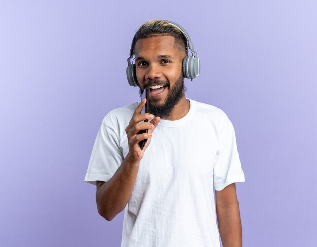 Joven afroamericano en camiseta blanca con auriculares sosteniendo smartphone usando como micrófono cantando feliz y alegre