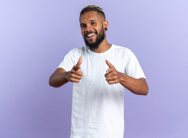 Joven afroamericano en camiseta blanca feliz y alegre mirando a la cámara sonriendo apuntando con el dedo índice a la cámara