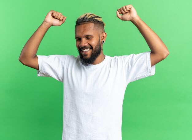 Joven afroamericano en camiseta blanca feliz y emocionado apretando los puños regocijándose de su éxito de pie sobre fondo verde