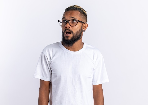 Joven afroamericano en camiseta blanca con gafas mirando a un lado confundido y sorprendido