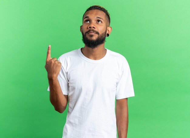 Joven afroamericano en camiseta blanca mirando hacia arriba con cara seria apuntando con el dedo índice a algo de pie sobre fondo verde