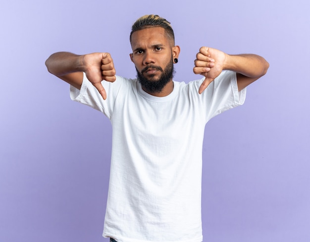 Joven afroamericano en camiseta blanca mirando a la cámara con cara seria apuntando con los dedos índices hacia abajo de pie sobre fondo azul.