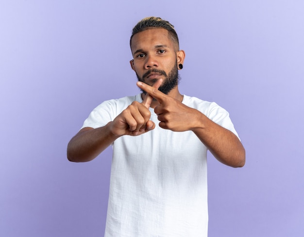 Joven afroamericano en camiseta blanca mirando a cámara con cara seria haciendo gesto de defensa cruzando los dedos índices de pie sobre fondo azul.