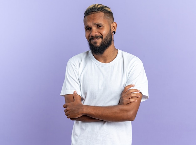 Foto gratuita joven afroamericano en camiseta blanca mirando a cámara confundido con los brazos cruzados